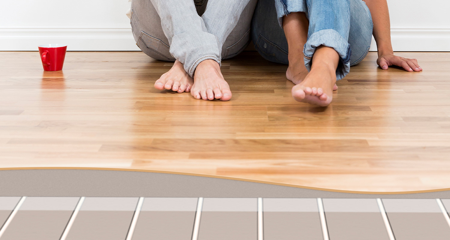 Två personer sitter barfota på ljust trägolv med röd mugg, underliggande golvvärmeskikt syns.