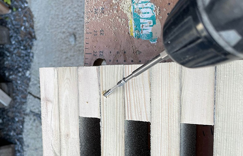 Borrmaskin skruvar en silverfärgad skruv i ljus träplanka med svarta skumlister på arbetsbänk.