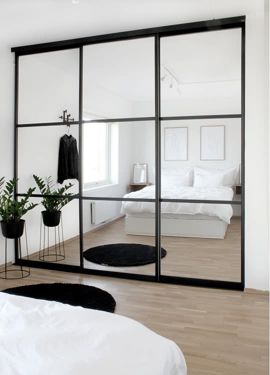 Vitt minimalistiskt sovrum med svart inramad glasvägg, vit säng, trägolv och gröna växter.