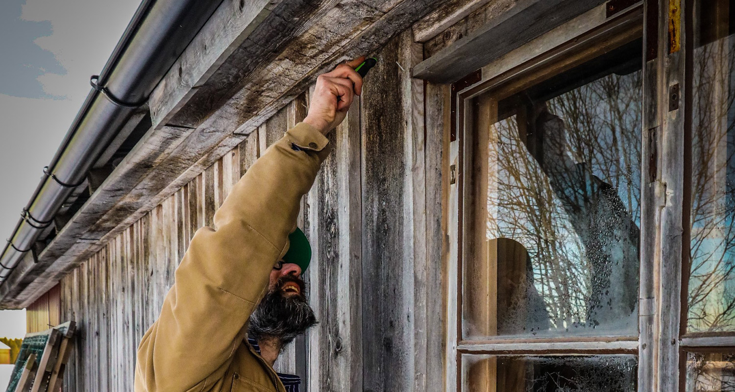 Man i beige jacka skruvar på väderbiten träfasad vid frostigt speglat fönster