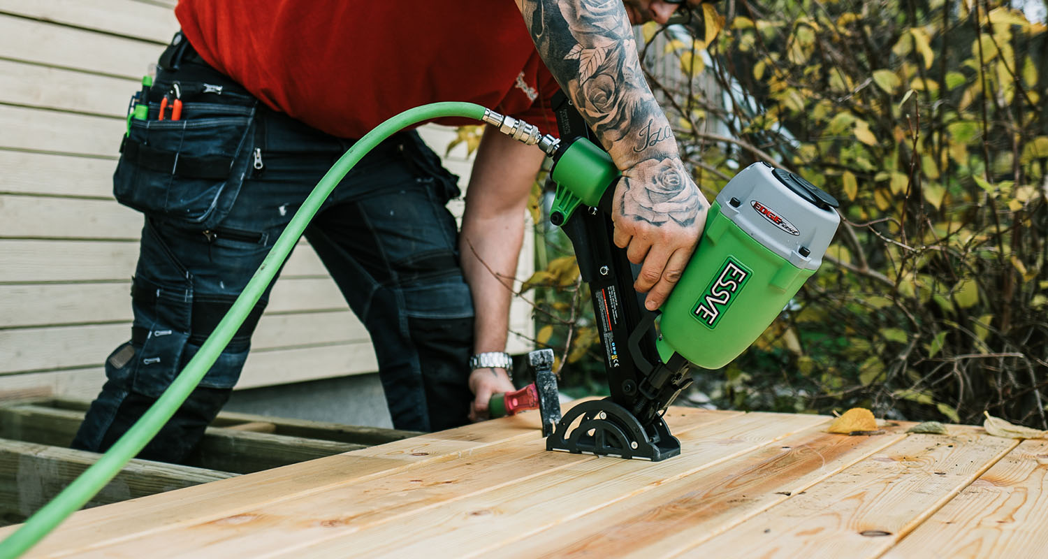 Snickare med tatuerad arm använder grön ESSVE-spikpistol på furubrädor utomhus