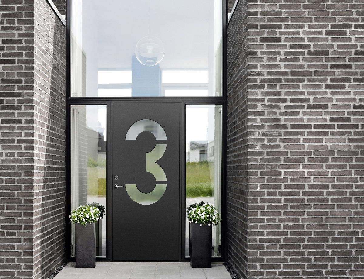 Svart ytterdörr med urfräst 3, smala glasfönster, tegelvägg och vita blomkrukor.