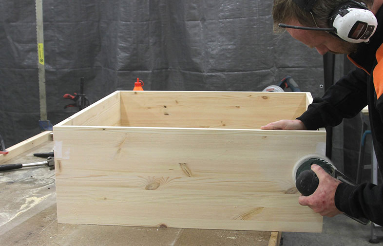 Person med hörselskydd slipar ljus träbox i verkstad med mörk bakgrund.