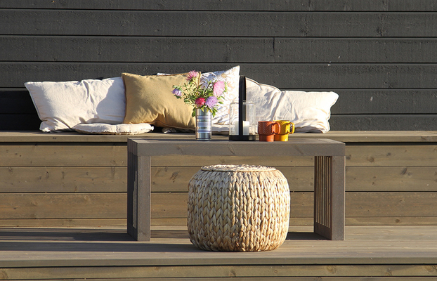 Uteplats med mörkgrå vägg, vita och blommiga kuddar, bord med blommor och muggar, rottingpuff