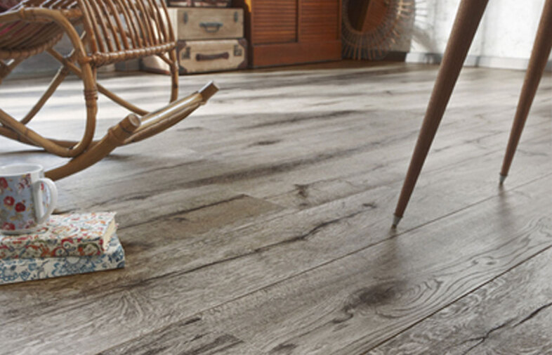 Gråbrunt trägolv med rottinggunga, blommig mugg på mönstrade böcker och möbelben.