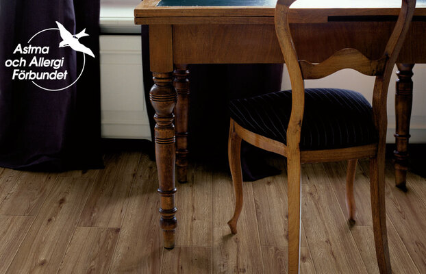 Brunt snidat skrivbord med stol på ljust trägolv, mörk gardin och Astma och Allergi Förbundet-logga.