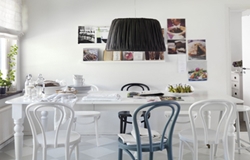 Ljust matrum med vitt bord, fyra vita och en svart stol under svart lampa, foton på vit vägg.