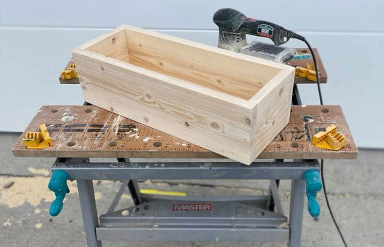 Rektangulär ljus trälåda på portabel arbetsbänk, med en svart-grön slipmaskin i bakgrunden.