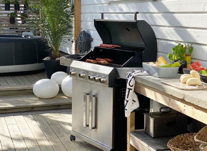 Silverfärgad gasolgrill med kött på trädäck bredvid bröd, sallad, majs och grönsaker