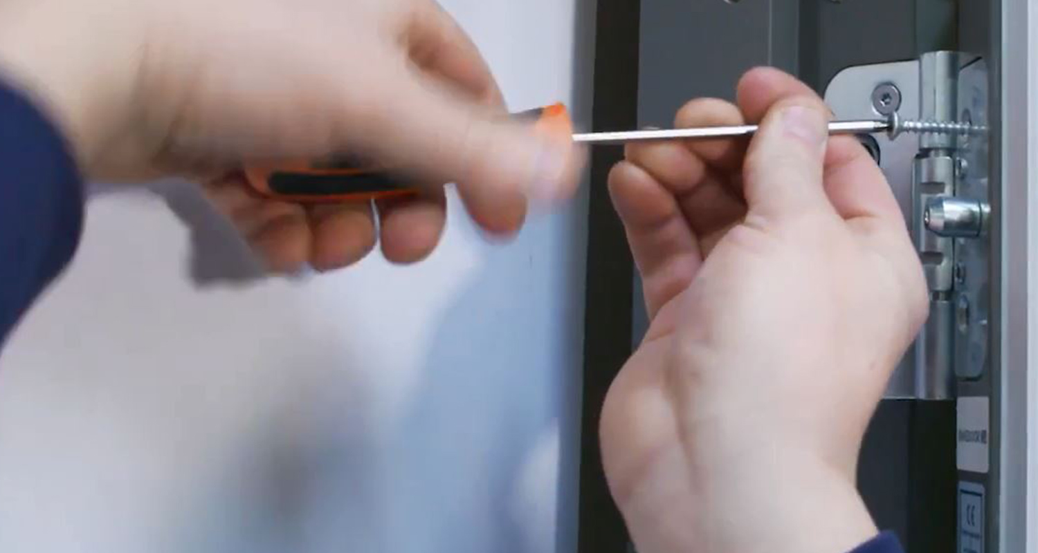 Närbild på händer som drar skruv i silverfärgat dörrgångjärn med orange-svart skruvmejsel