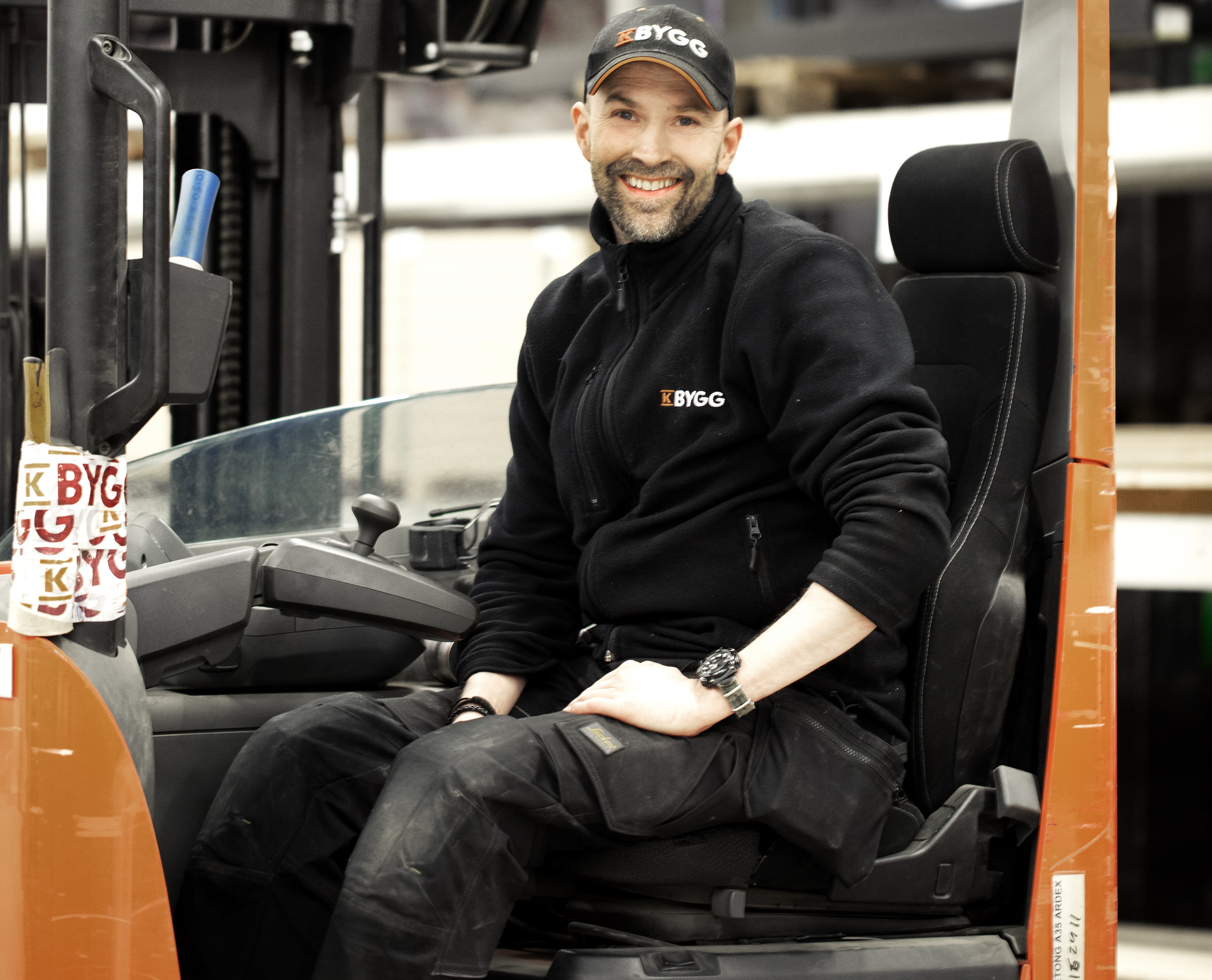 Arbetsman i svart BYGG-uniform och keps sitter i orange truck i lager.