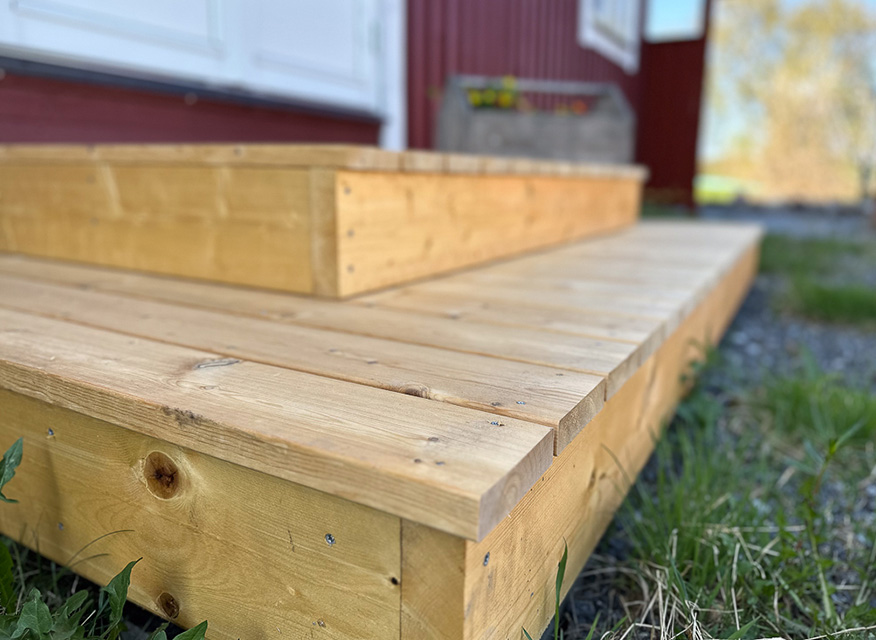 en nybyggd trappa i trä med två steg, placerad utomhus framför en byggnad med röd fasad. Träets struktur syns tydligt och runt trappan finns gräs och grus.
