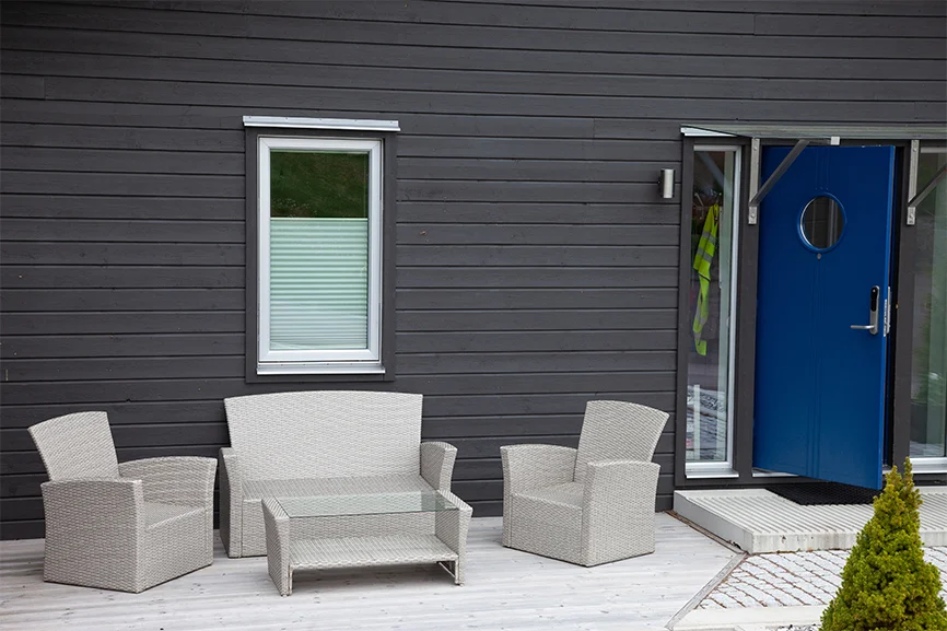 Mörkgrå träfasad med vitt rottingmöblemang på ljus altan framför blå dörr och fönster