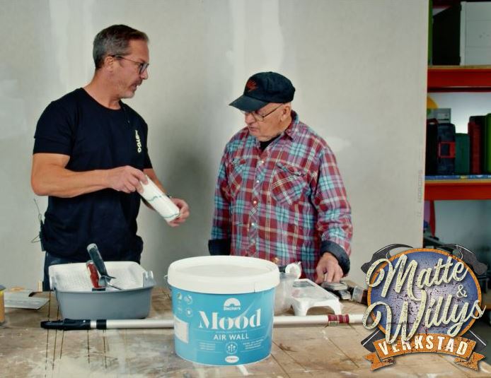 Två hantverkare i verkstad vid bord med blå-vit färgburk, roller och målartillbehör.