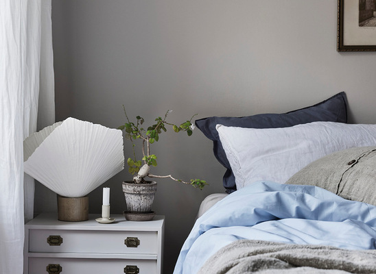 Sängbord med vit vikbar lampa, miniatyrträd i kruka och ljus, bredvid ljusblå bäddning.