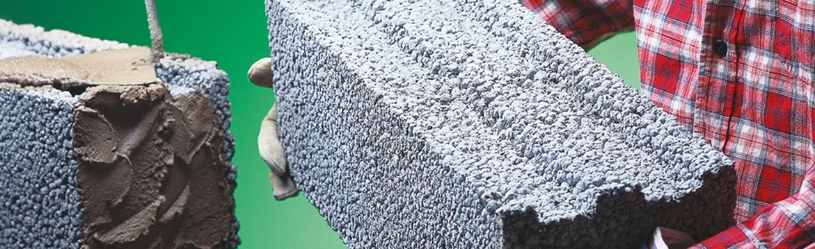 Handskbeklädda händer håller grå porös betongsten med murbruk, röd rutad skjorta mot grön bakgrund