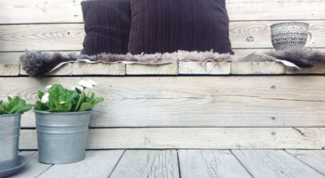 Sittbänk i ljust trä med grått fårskinn, mörkgrå kuddar, kopp och zinkkrukor med vita blommor