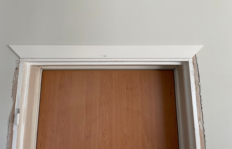 Övre del av ljust träinomhusdörr med vit karm i vitmålad vägg, små skador runt fogen
