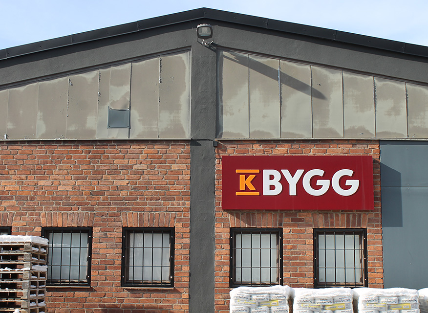 Röd KBYGG-skylt på tegelvägg med grå vindskiva, svarta gallerfönster och snötäckta säckar.