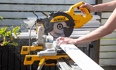 Händer håller vit planka mot gul DeWalt-kapmaskin utomhus, sågspån sprutar.