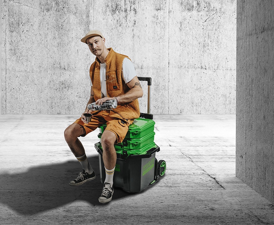 Man i orange kläder sitter på grön-svart verktygsvagn och häller dryck i mugg mot grå betongvägg.