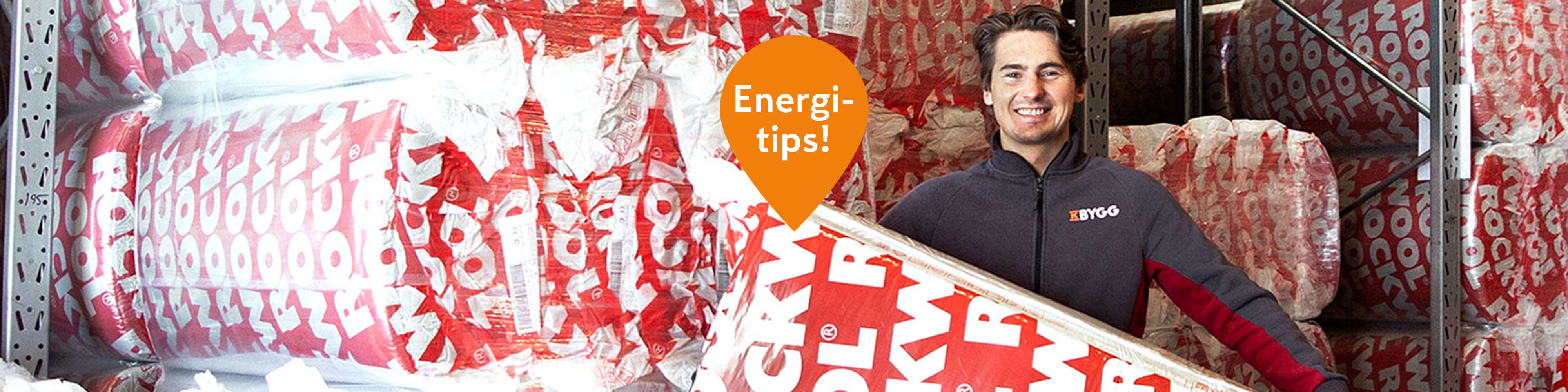 Blågråklädd anställd håller isolering framför hylla med rödvita rullar och orange Energi-tips