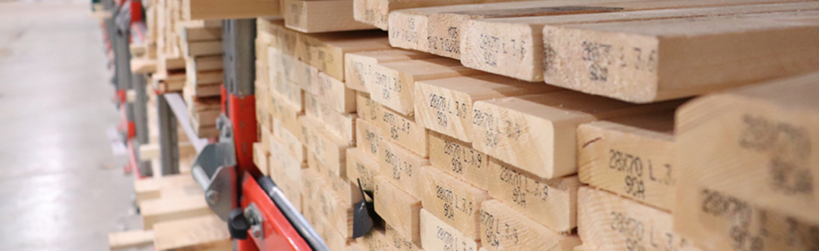 Staplad ljusbrun trälast på röd-grå metallhylla i lager.