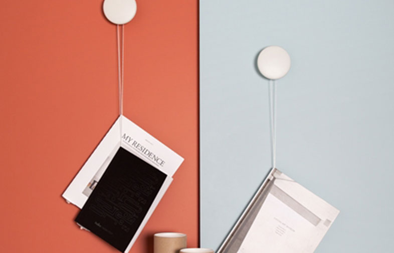 Två vita cirkulära väggkrokar med snören håller tidningar mot terrakotta- och ljusblå vägg.