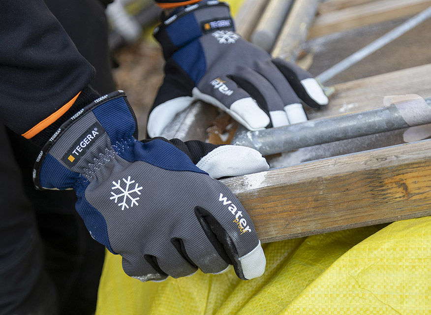 Ett par vinteranpassade arbetshandskar i grått och blått med vita detaljer. Handskarna har snöflinga-symbol och texten water, vilket indikerar kyltåliga och vattenavvisande egenskaper. Märket TEGERA syns vid handleden.