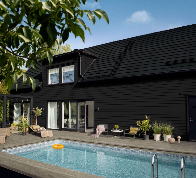 Svart trävilla med glasdörrar, pool med ljusblått vatten och orange badring, utemöbler och växter