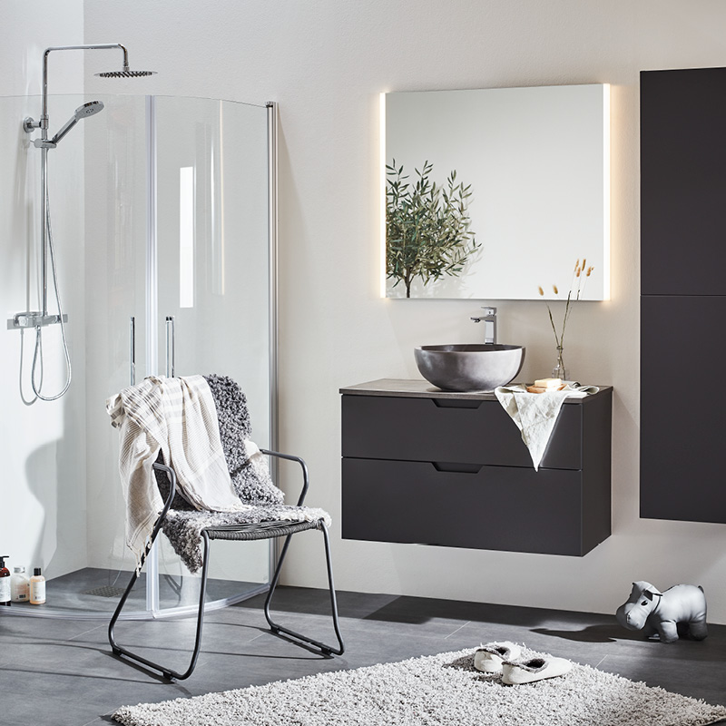 Modernt badrum med glasdusch, svart flytande kommod med runt handfat, LED-spegel och stol.