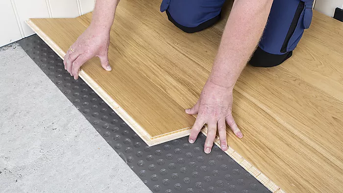 Händer som lägger ljus träplanka på svart bubbelmatta över grå betong.