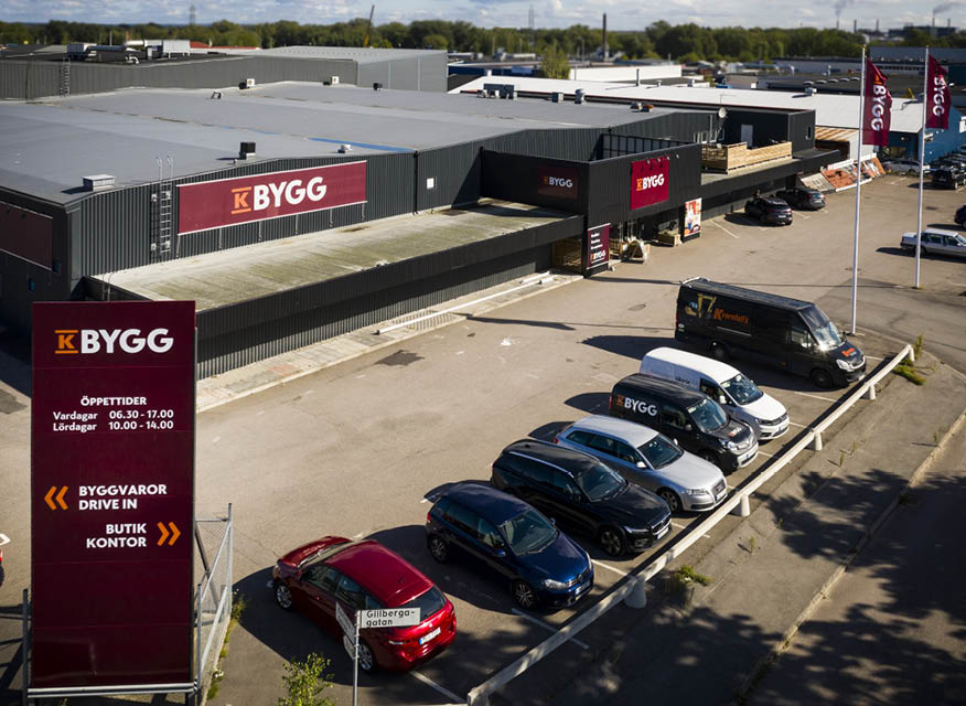 Flygfoto över grå byggvaruhus "K Bygg" med röda skyltar, bilar och skylt med öppettider.