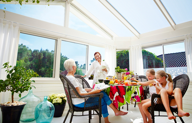 Soligt uterum med vitt snedtak, blommig duk, frukostbord med juice, växter och familj.