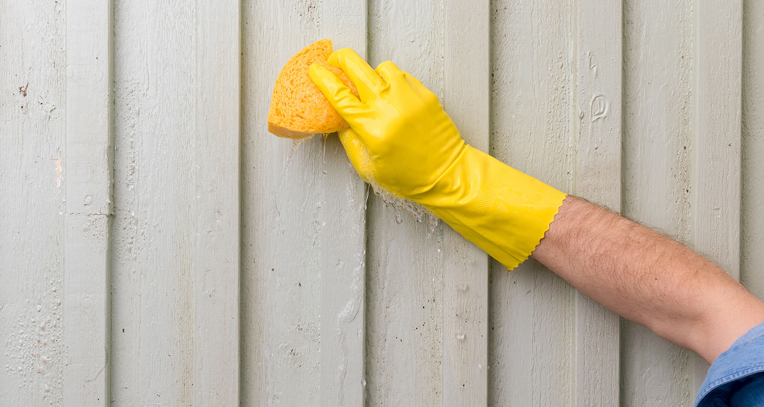 Hand i gul gummihandske torkar grå träpanel med gul svamp, vattendroppar