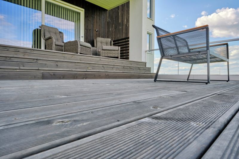 Modern träterrass med gråbruna plankor, rottingfåtöljer, metallbänk och glasräcke mot blå himmel