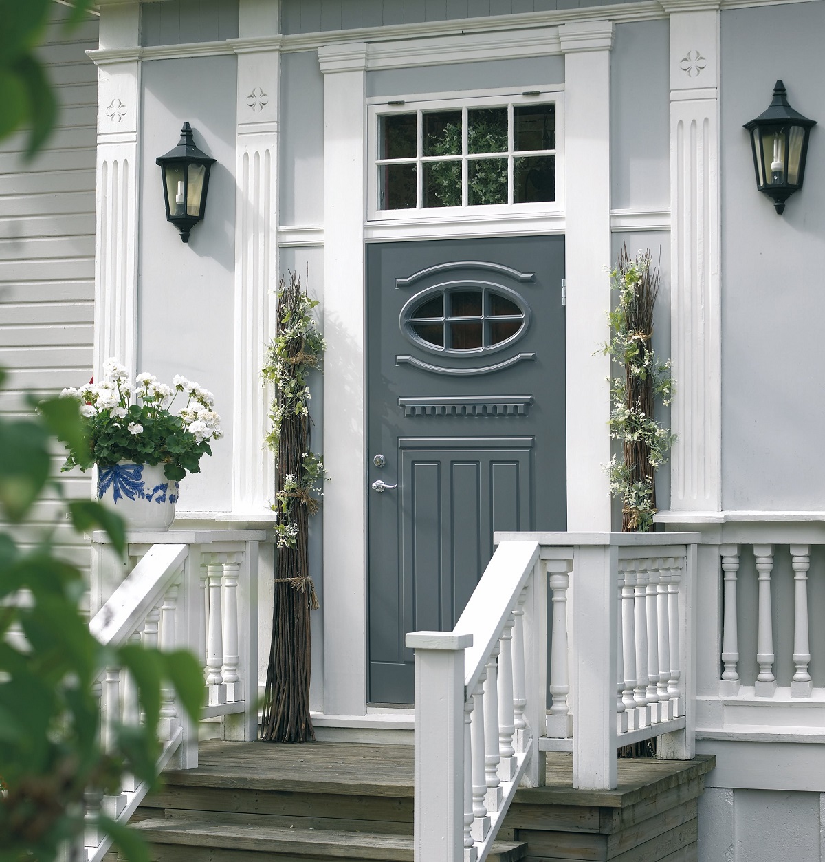 Vit träfasad med grå ytterdörr med ovalt fönster, svarta lyktor och blommor i blåvit kruka