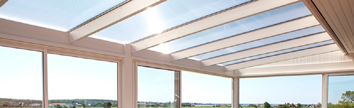 Inglasad vit veranda med glasbjälkar i taket, blå himmel och gröna fält utanför