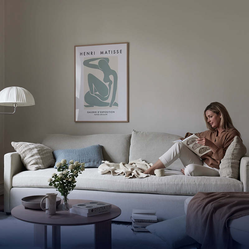Ljust vardagsrum med beige soffa där en person läser, Henri Matisse-affisch, kaffebord med blommor.