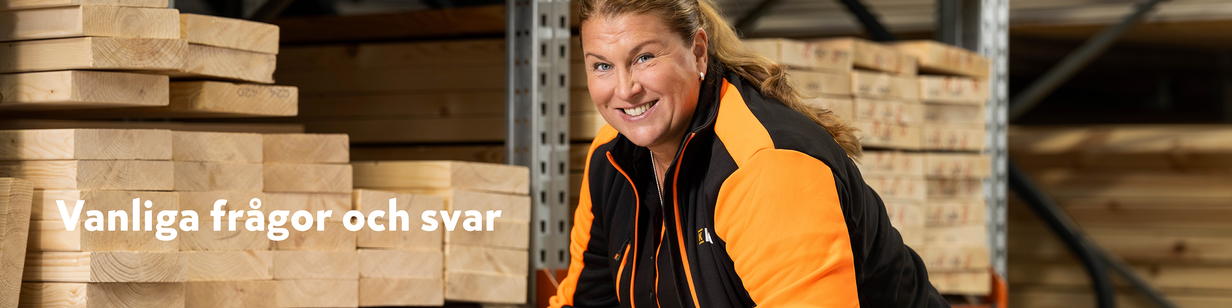 Stapel ljusa träplankor i lagermiljö med person i orange-svart arbetsjacka.