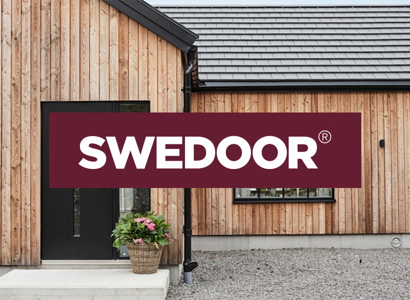 visar en modern husfasad i träpanel med en mörk ytterdörr till vänster. Framför dörren står en korg med blommor. Ovanpå motivet ligger en vinröd banderoll med den vita logotypen SWEDOOR.