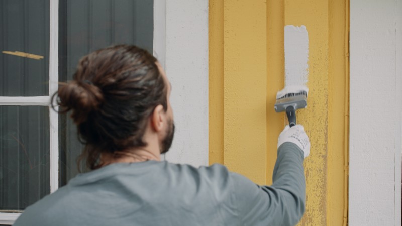 Man med knut målar gul fasadpanel vit med pensel, bär handskar och grå tröja framför fönster.
