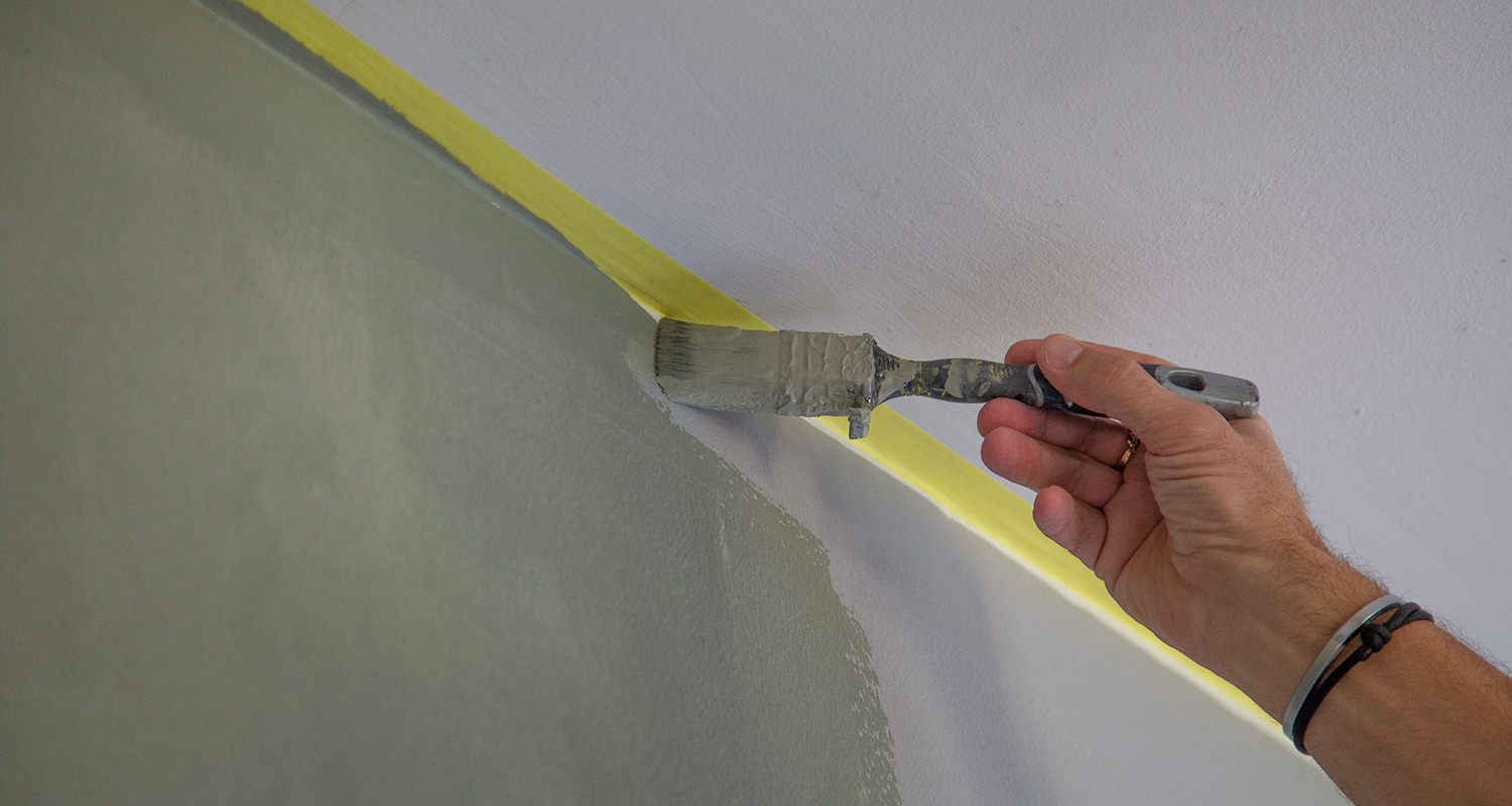 Hand med pensel målar grågrön vägg vid vit takkant med gul maskeringstejp