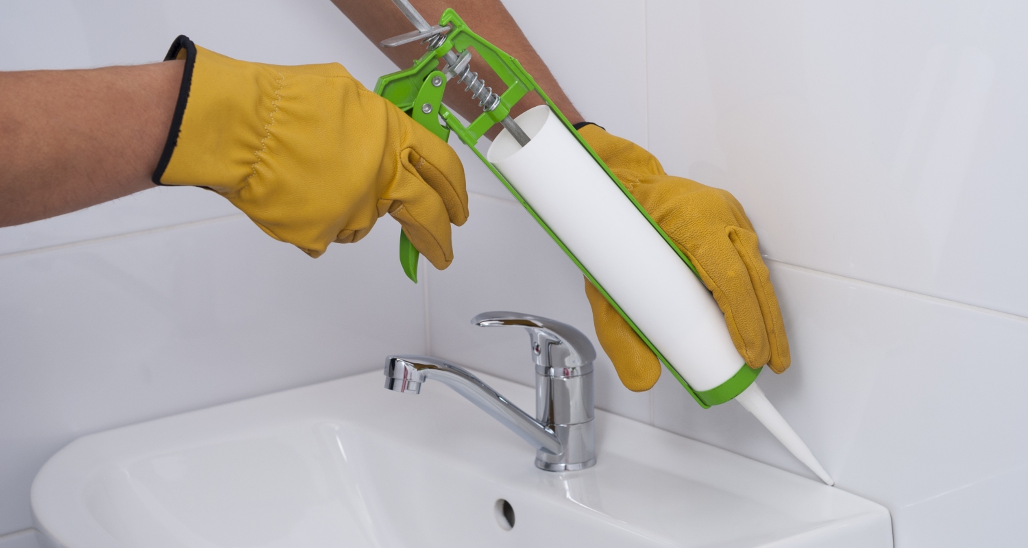 Gula handskar håller grön fogpistol för vit fogmassa vid vit handfat och kakel