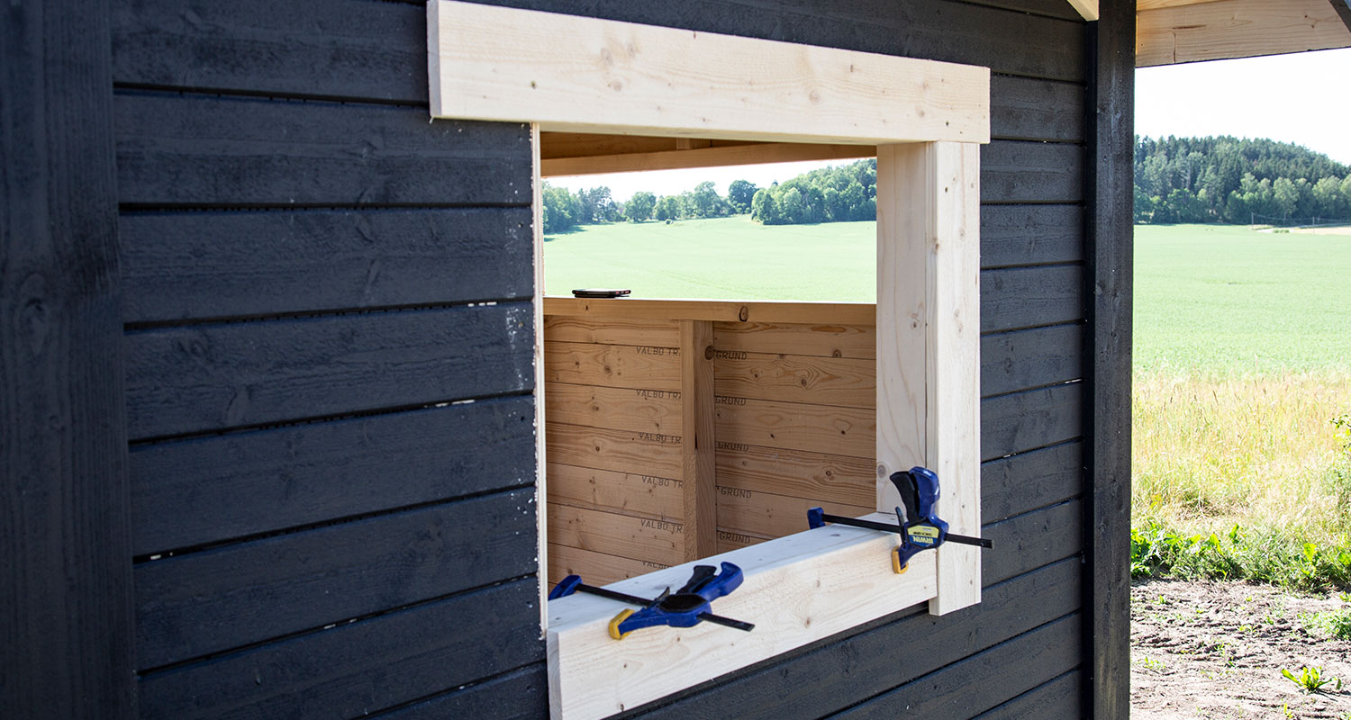 Omålat träfönster i svart trävägg med blå-gula klämmor, utsikt över grönt fält