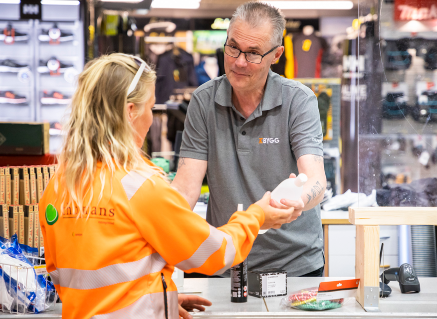 Butiksmedarbetare i grå pikétröja överlämnar plastflaska vid kassa till kund i orange skyddsjacka.