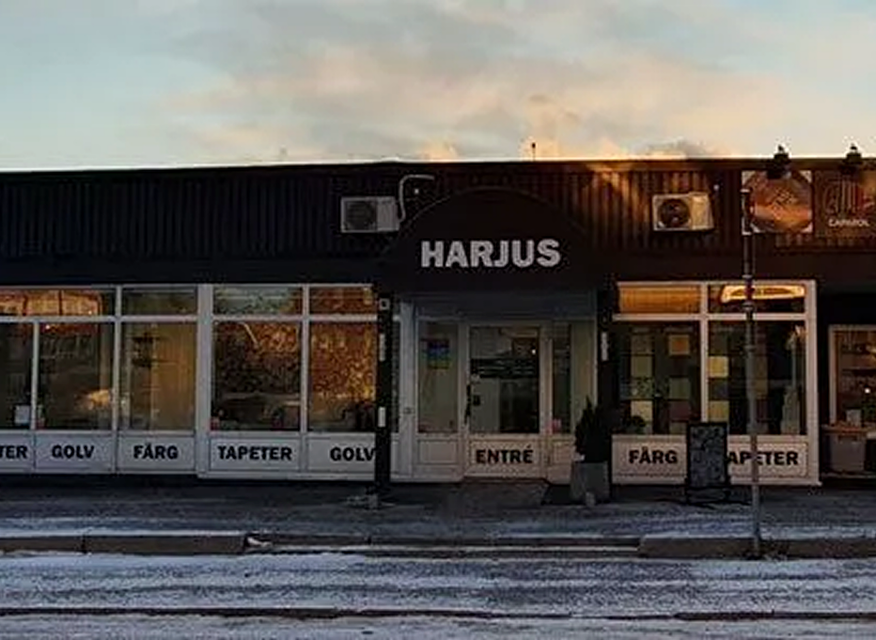 En butik med svart fasad och stora glasfönster, där skylten ovanför entrén visar namnet HARJUS och fönstren annonserar produkter som golv, färg och tapeter.