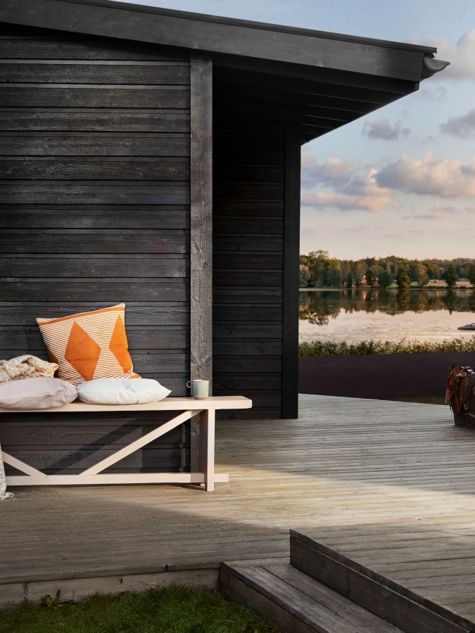 Trädäck vid svart trästuga med bänk, orange/vit kudde och sjöutsikt i kvällsljus