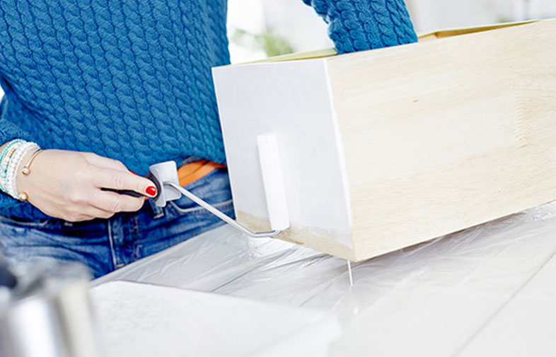 Hand med röda naglar och blå tröja målar trälåda vit med liten roller på plastskydd