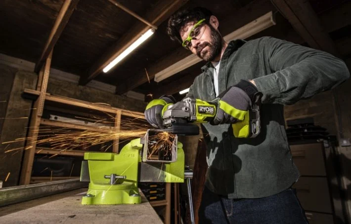 Person slipar metall i grönt skruvstäd med grön Ryobi-vinkelslip, gnistor flyger i mörkt garage.